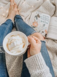 Eine Person sitzt auf einer Decke und hält eine Tasse Cappucchino in der Hand, im Hintergrund liegt eine aufgeschlagene Bibel